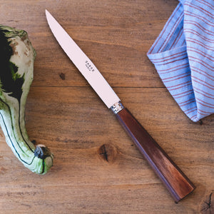 The Sabre Nature Steak Knife, featuring a wood handle and stainless steel blade, rests on a wooden surface beside a striped cloth and a green vegetable.