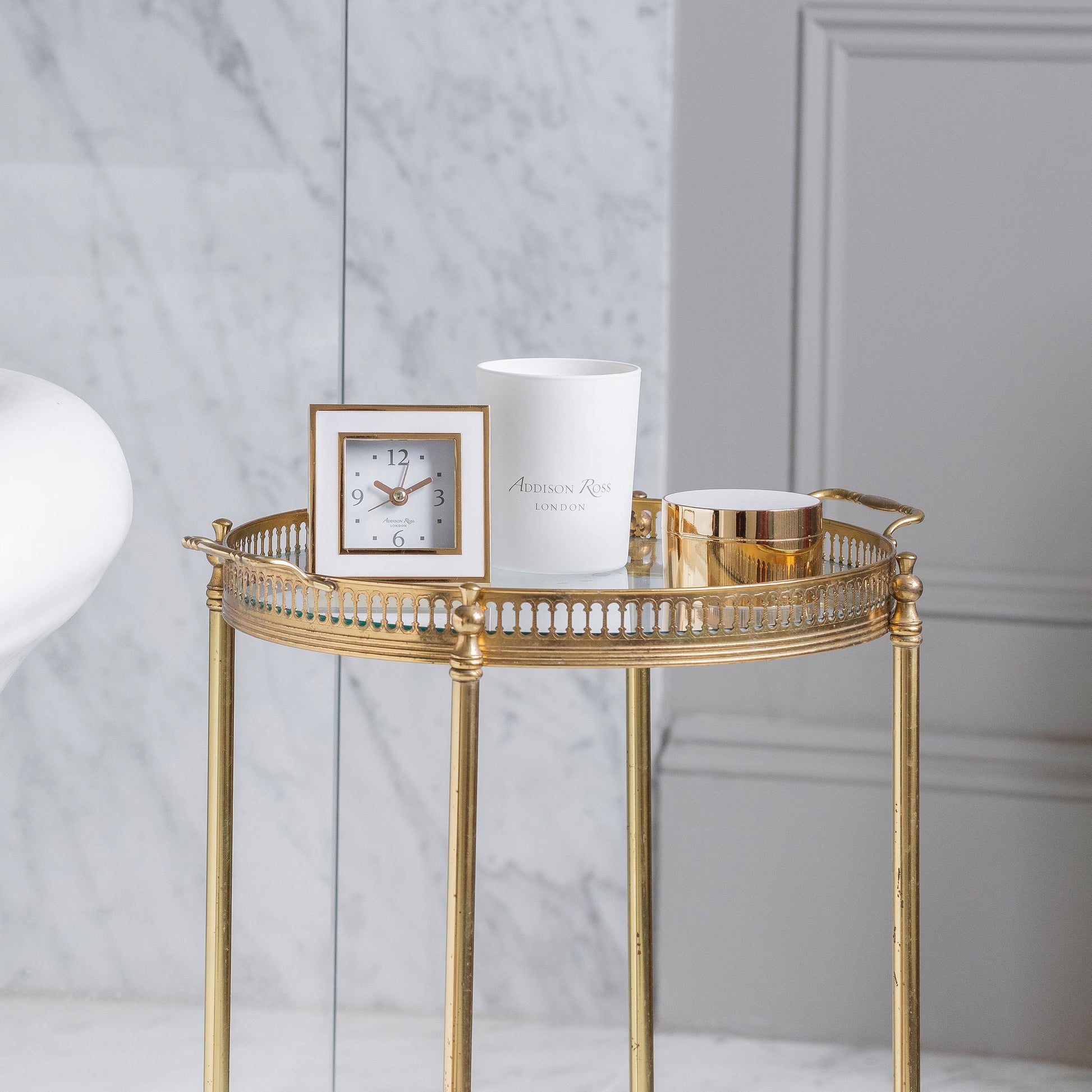 A gold side table features a white candle, an Addison Ross Gold Square Alarm Clock with silent sweep movement, and a round gold container against a marble wall backdrop.