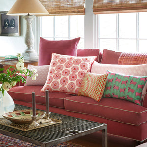 A living room with a pink sofa, patterned throw pillows including the John Robshaw Vikas Lotus Bolster, a metal coffee table, candles, a floral vase, and large windows with bamboo blinds in the background.