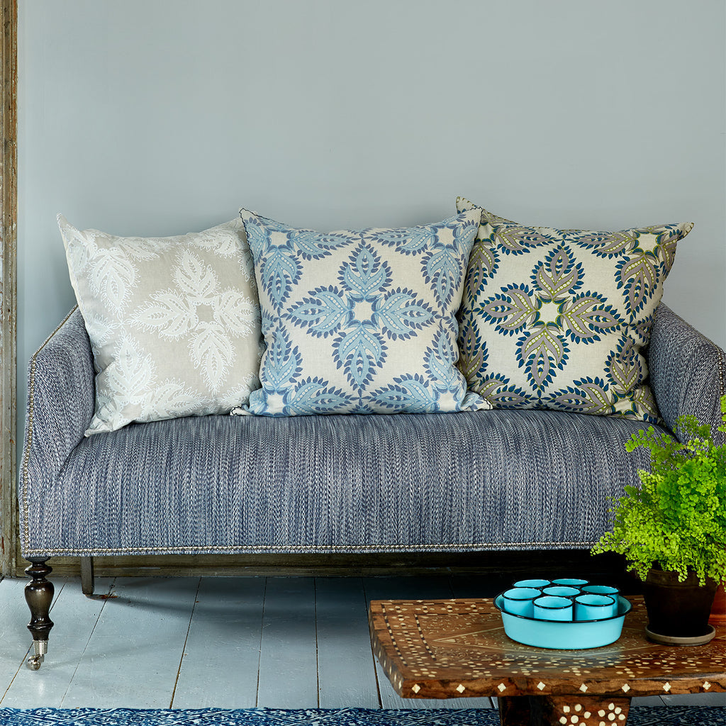 A blue upholstered sofa in a cotton-linen blend features three John Robshaw Verdin Euro Pillows in neutral and blue, beside a wooden table with a potted fern and a tray of blue bowls.