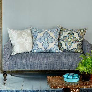 A blue upholstered sofa in a cotton-linen blend features three John Robshaw Verdin Euro Pillows in neutral and blue, beside a wooden table with a potted fern and a tray of blue bowls.