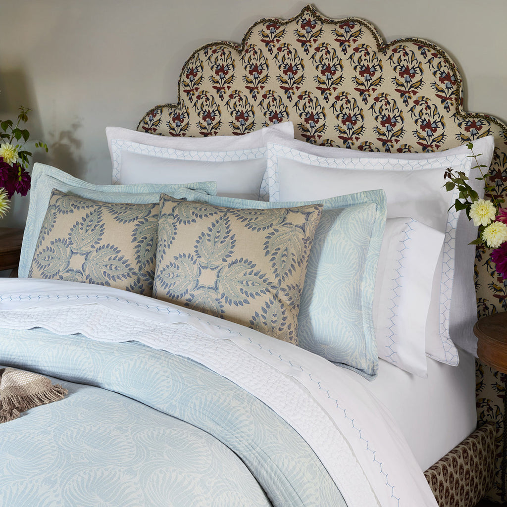 A neatly made bed with cotton linen bedding, the John Robshaw Verdin Lapis Pillow, a decorative upholstered headboard, and flowers on bedside tables.