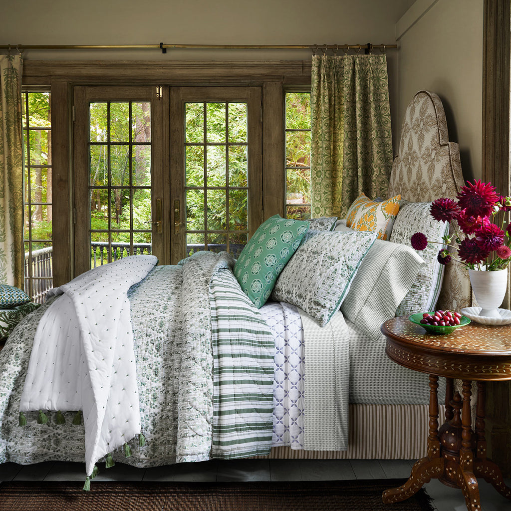 A cozy bedroom featuring the John Robshaw Lina Sage Bedding Collection, adorned with floral decorations. French doors open to a green garden, while soft cotton voile curtains add an airy touch.