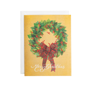 Caspari's Leaf Wreath with Plaid Bow Boxed Card Set features a festive wreath with red berries and a plaid bow, "Merry Christmas" below on a gold background. An envelope is partially visible behind the card.