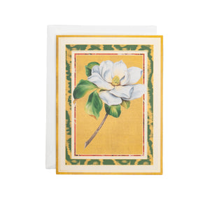 The Magnolia Bloom Boxed Card Set by Caspari features a white magnolia with green leaves on a yellow background, bordered in green and gold, and comes with a matching white envelope.