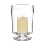 A lit cream pillar candle sits inside a clear glass hurricane holder with a pedestal base from the Nantucket Hurricane Collection by Simon Pearce, evoking the look of their classic trifle dish.