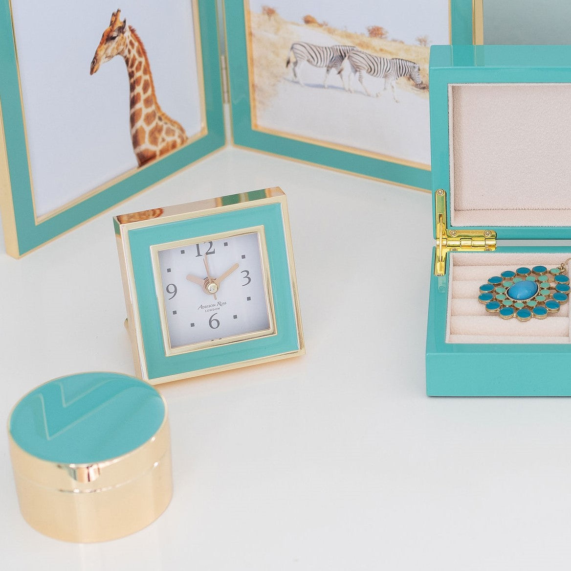 An Addison Ross Gold Square Alarm Clock showcases its turquoise and gold enamel finish with silent sweep movement next to a jewelry box containing a blue necklace. Framed giraffe and zebra photos complement the circular container on the luxurious velvet backdrop.