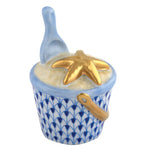 The Herend Beach Bucket Collection features a hand-painted white porcelain bucket with blue patterns, filled with white material, topped with a gold starfish, and includes a blue scoop inside.