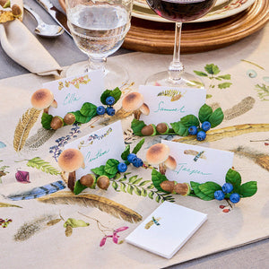 Four place card holders from the Juliska Forest Walk Tabletop Collection, adorned with faux mushrooms, acorns, leaves, and berries, are displayed on a botanical tablecloth next to glasses of water and red wine.