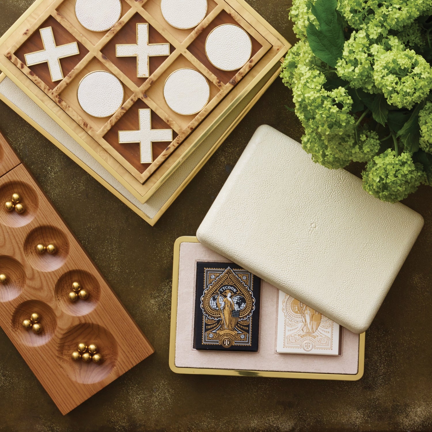 The Aerin Shagreen Card Set in Cream, featuring two decks in a gold-embossed box, sits alongside a tic-tac-toe board, wooden marble game, and green hydrangeas on a dark surface.
