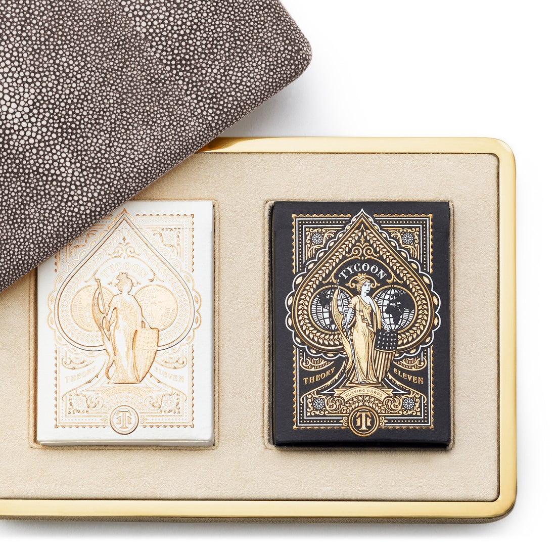 The Aerin Chocolate Shagreen Card Set features two ornate playing card decks—one white and one black with gold details—displayed side by side on a beige tray next to a partially visible chocolate shagreen card case with brass accents.