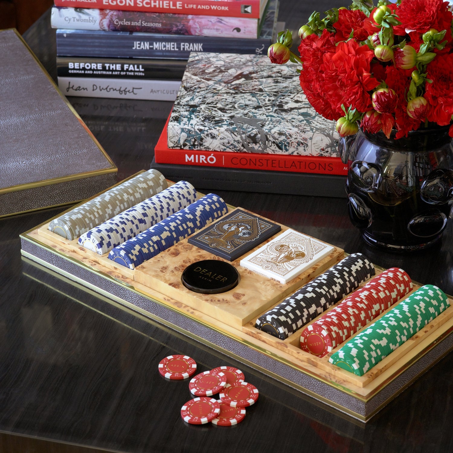 The Shagreen Poker Set in Chocolate by Aerin—with poker chips, playing cards, and a dealer button—rests beside red flowers and art books in its luxurious case.