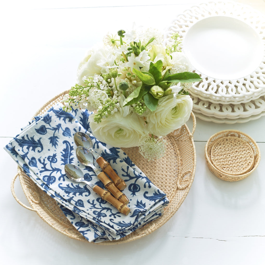 The Aerin Raffia Round Tray displays blue and white napkins with bamboo-handled spoons next to a white floral arrangement. White decorative plates, a woven coaster, and coastal-inspired accents enhance the elegant setting.