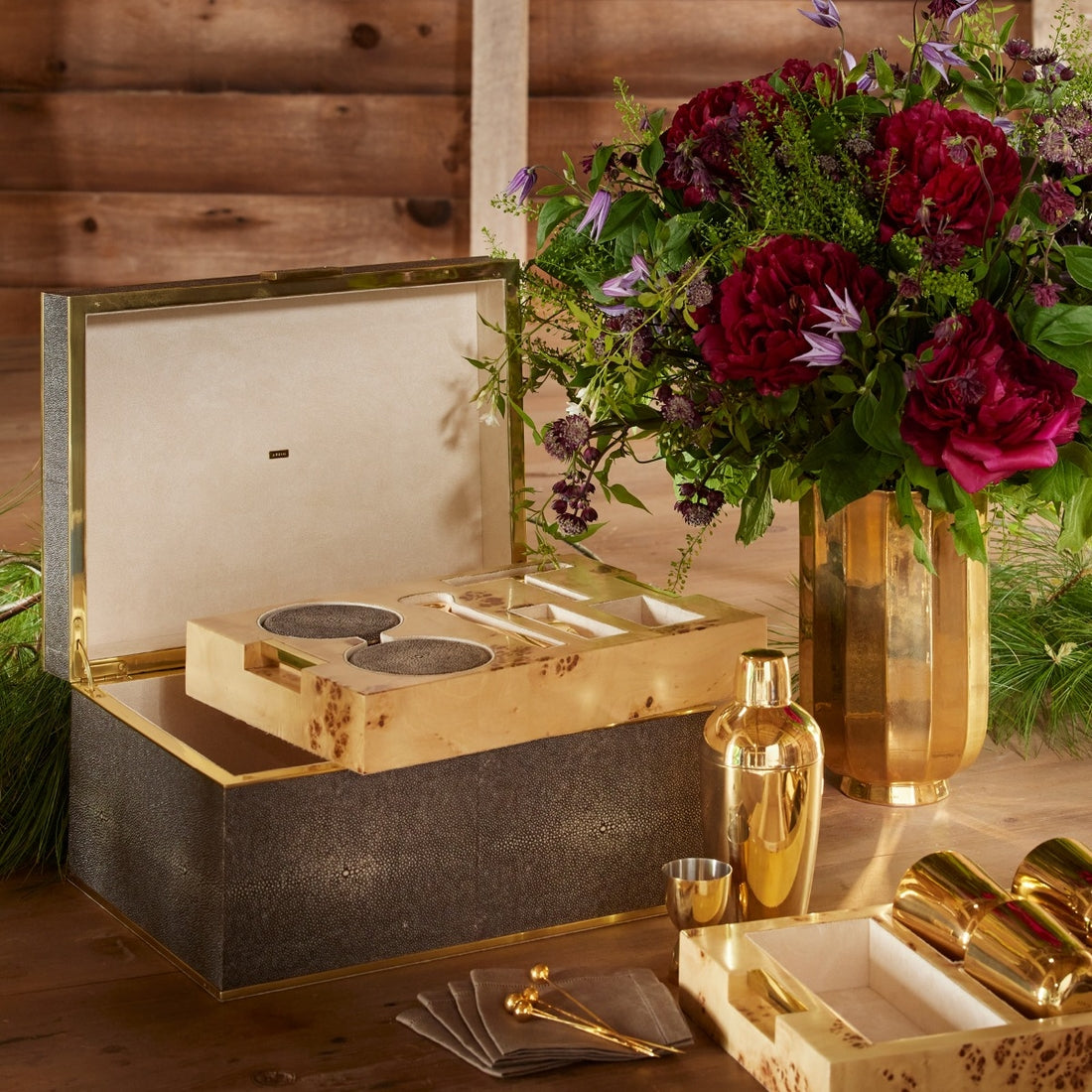 The Chocolate Classic Shagreen Bar Set by Aerin, featuring cocktail accessories, sits beside a gold vase with red and purple flowers on a wooden table—an elegant home bar set display.