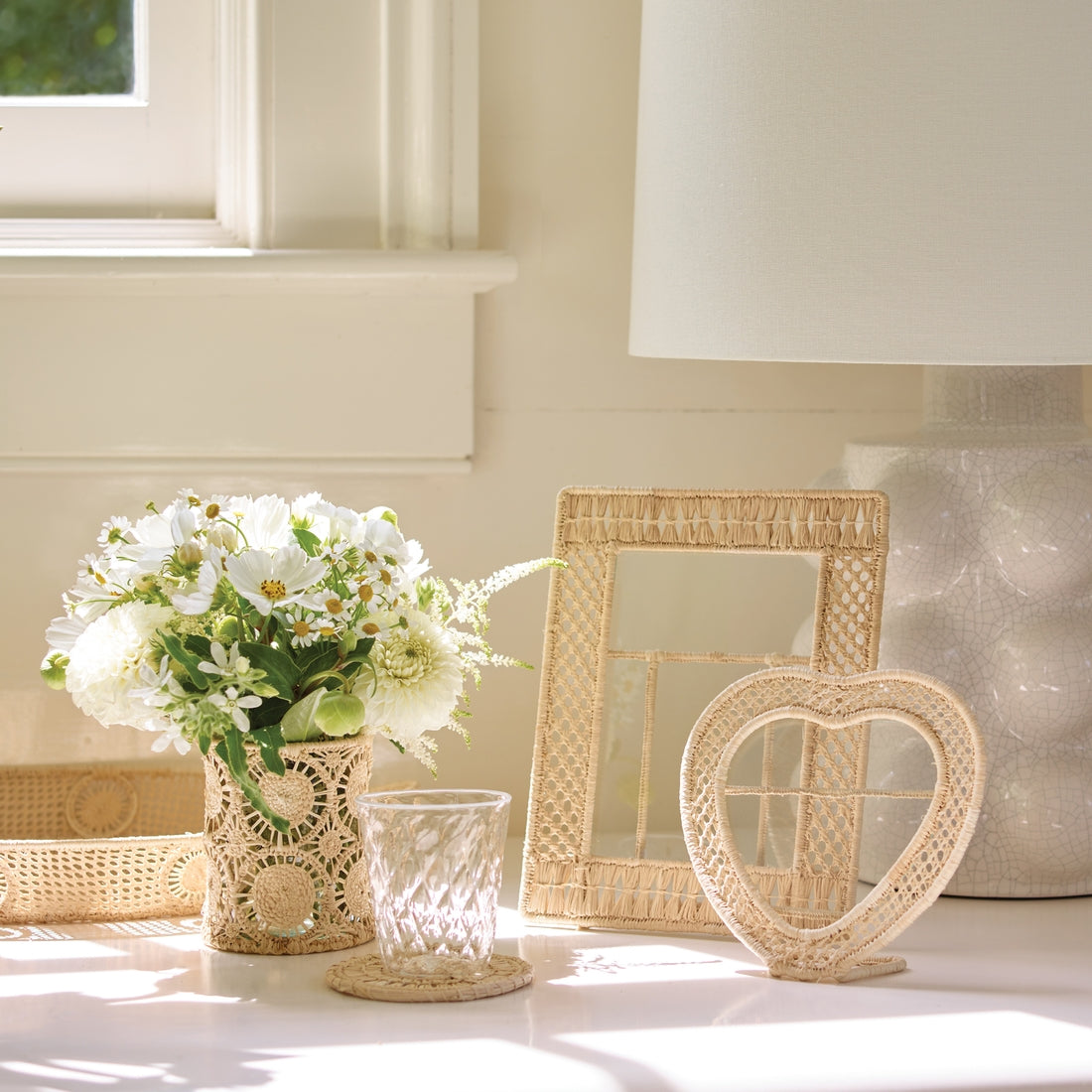 A white tabletop displays a glass of water, a vase with white flowers, the Aerin Raffia Frame for organic appeal, a lamp, and round coasters near a sunlit window.