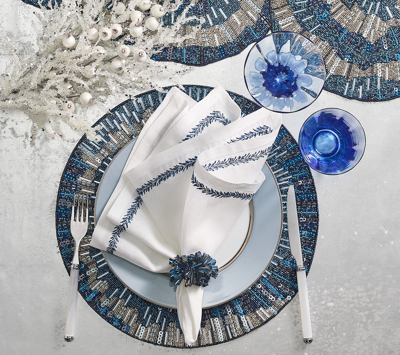 A table setting with a white napkin embroidered with blue leaves, blue glassware, silverware, a Kim Seybert Illusion Placemat, and a decorative white branch centerpiece.