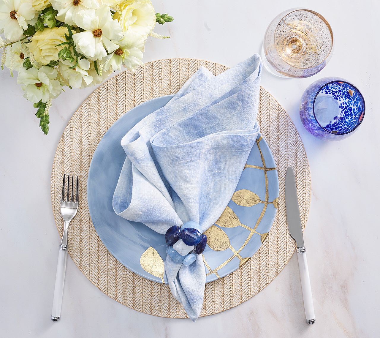 Create a fresh spring and summer table with a light blue napkin on a blue plate, Kim Seybert Herringbone Placemat in polyvinyl with gold accents, fork, knife, two glasses, and a white floral centerpiece.