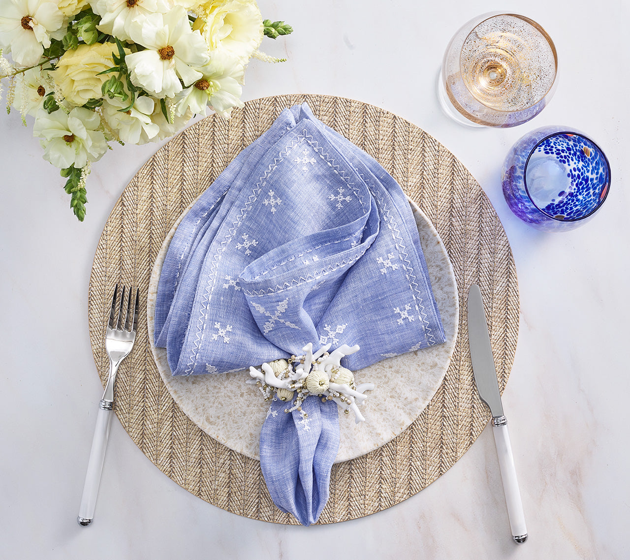A fresh spring table set with a white plate, blue embroidered napkin, silverware, clear and blue glasses, and a floral arrangement on the Kim Seybert Herringbone Placemat—perfect for seasonal dining.