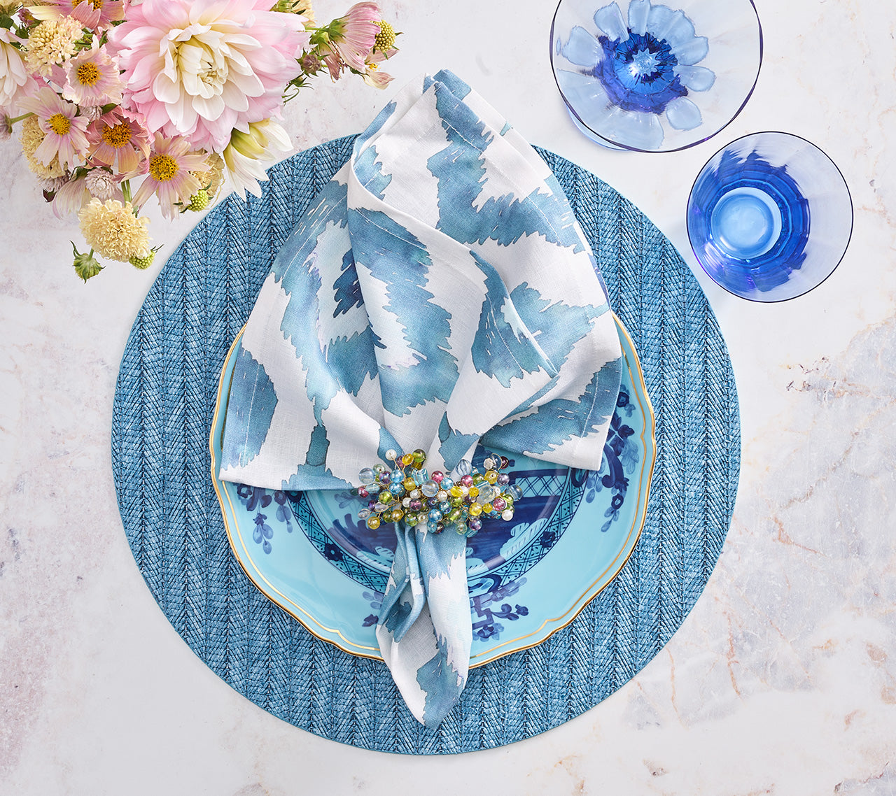 A blue and white table setting features a patterned napkin, floral napkin ring, blue plates, Kim Seybert Herringbone Placemat, blue glassware, and a floral bouquet on marble—perfect for spring and summer dining.