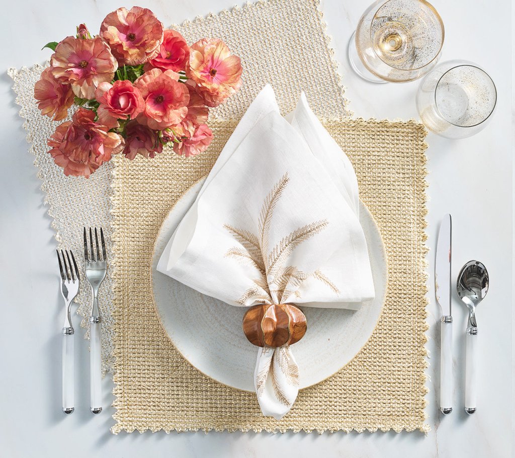 A table setting with a white plate, Kim Seybert Silver & Gold Stamped Reversible Placemat, folded white napkin in a holder, silver cutlery, two glasses, and a vase of pink flowers.