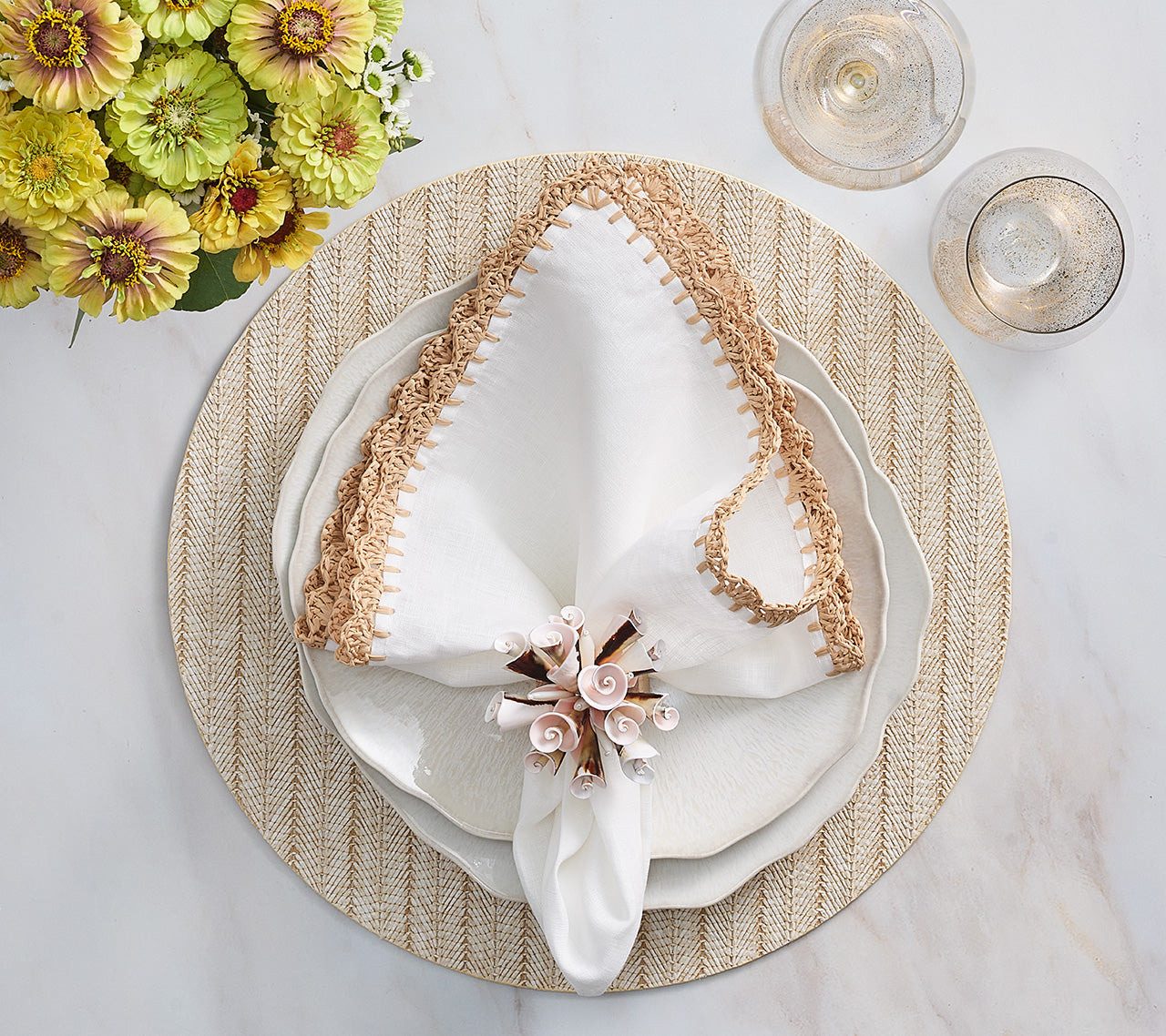 Table setting with a white plate, white napkin with tan trim held by the Kim Seybert Seashell Burst Napkin Ring, round woven placemat, two sparkling drinks, and a vase of yellow flowers—ideal for seaside gatherings.