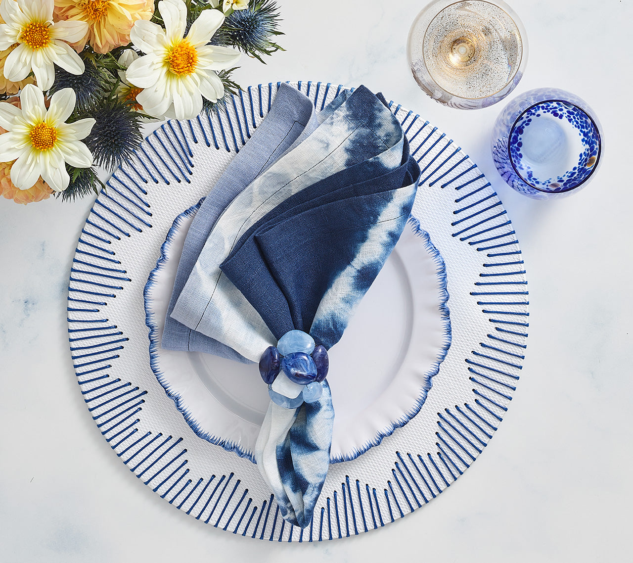 A blue and white tie-dye napkin with a blue ring rests on a patterned plate set atop the Kim Seybert Dream Weaver Placemat, beside a drink, a blue glass, and bright summer flowers—ideal for stylish entertaining.