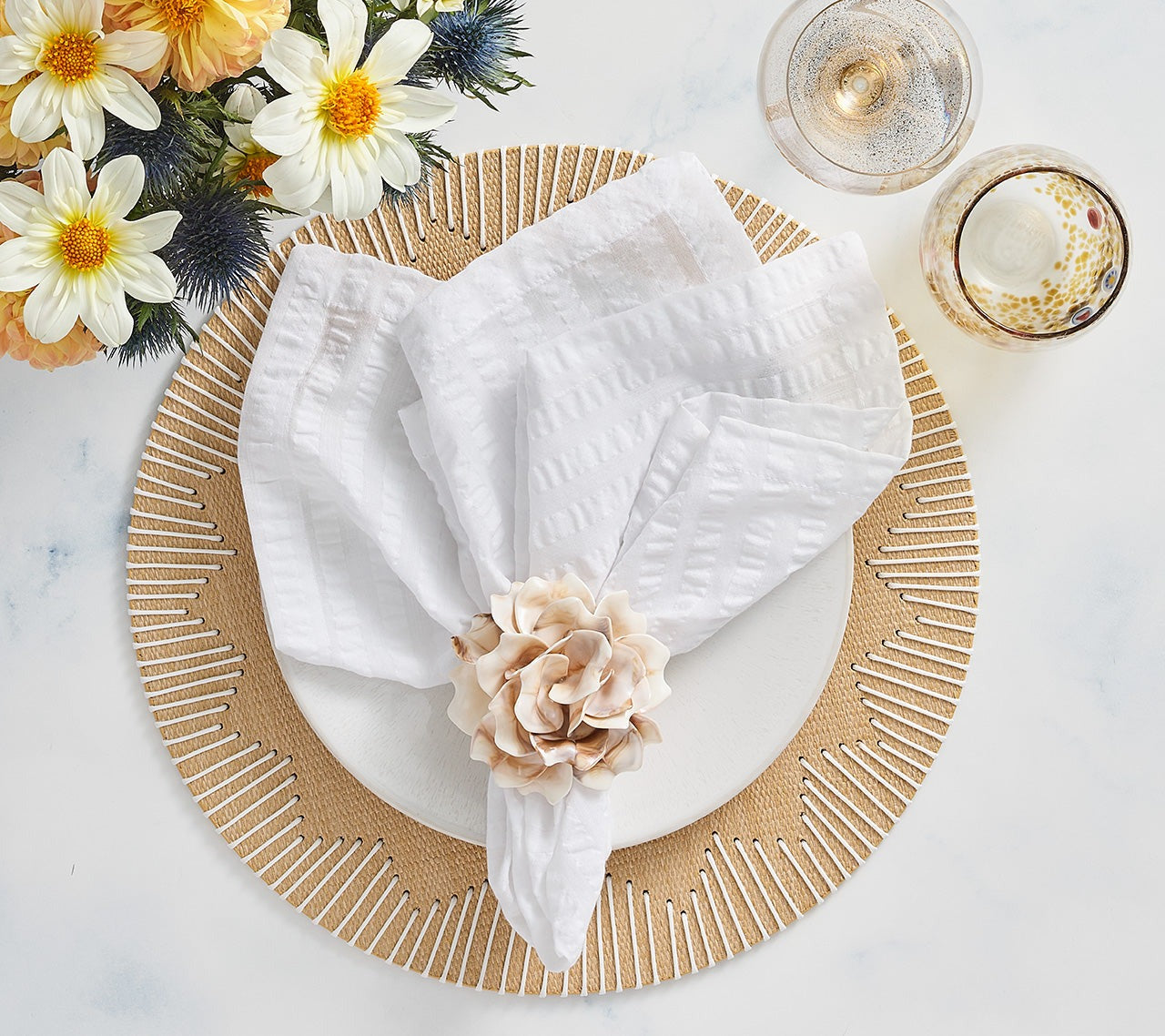 A white plate with a textured napkin and a beige flower napkin ring rests on a Kim Seybert Dream Weaver Placemat, next to two drinks and a vase of white and yellow flowers—ideal for summer entertaining.