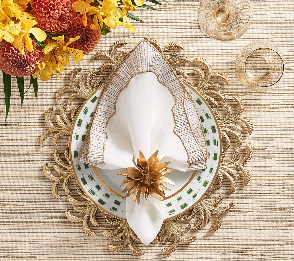 A formal table is set with a decorative gold placemat, white plate with green accents, Kim Seybert's Frame Napkin Set folded with gold trim and floral ring, metallic touches, two glasses, and a yellow-red floral centerpiece.