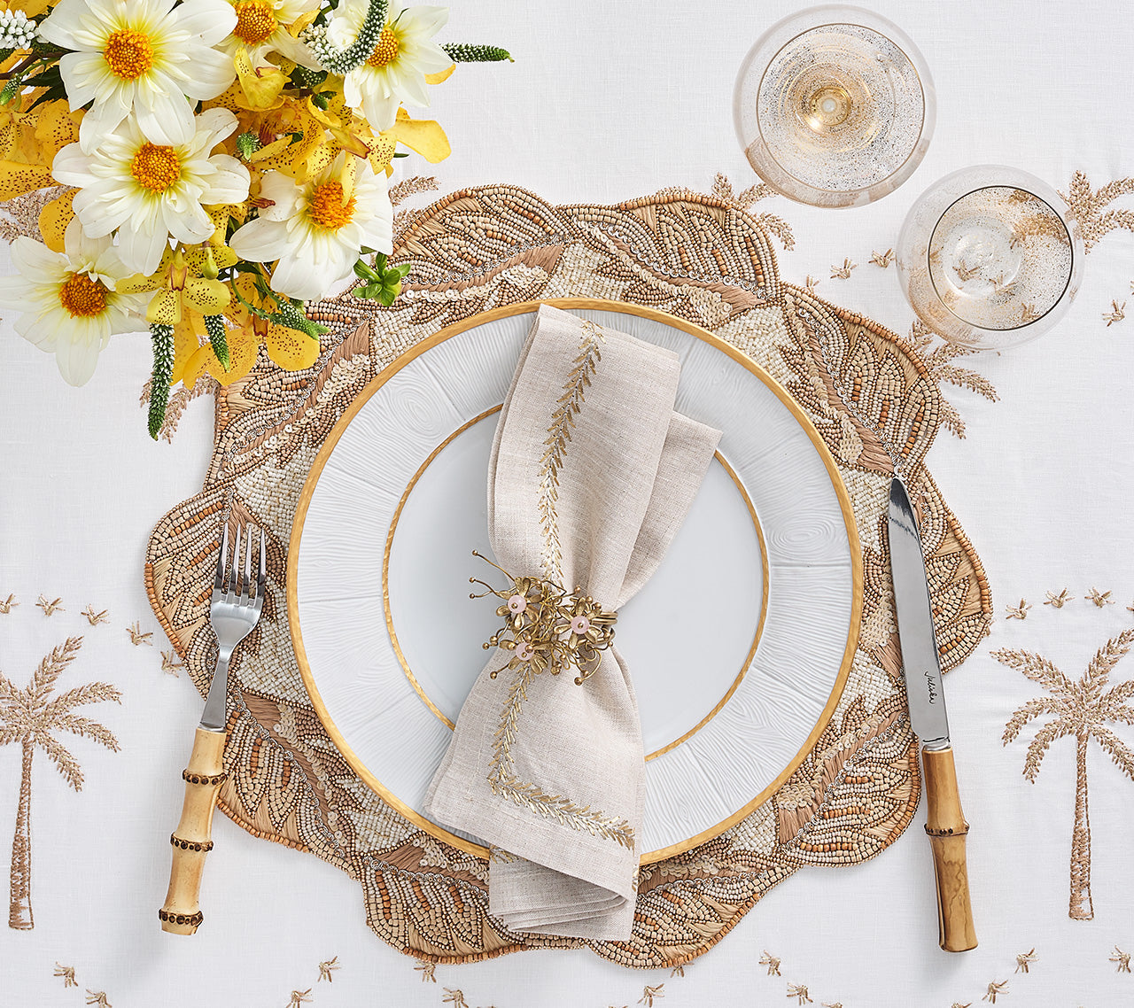 A table setting with a white plate, gold-edged napkin, floral napkin ring, bamboo cutlery, two glasses, yellow and white flowers on an embroidered cloth is completed with the Kim Seybert Winding Vines Placemat in natural tones.