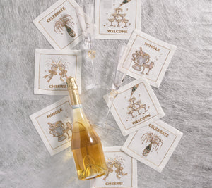 A bottle of champagne, two glasses, and the Kim Seybert Cheers Cocktail Napkin Set featuring six embroidered linen napkins are arranged on a silvery textured surface.