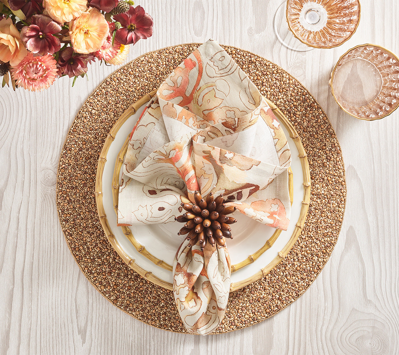 A table setting featuring Kim Seybert’s Giverny Napkin Set with a brown napkin ring, a gold beaded charger, white plate, and crystal glassware on light wood, accented by flowers.