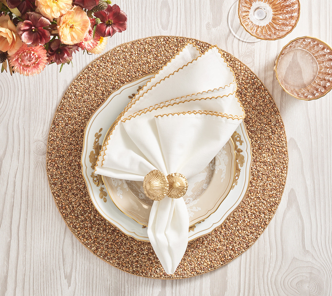 Table setting featuring a gold and white plate, white napkin with gold trim and shell-shaped ring, gold-toned glasses, flowers, all atop the Kim Seybert Emberfall Placemat on a light wood table.