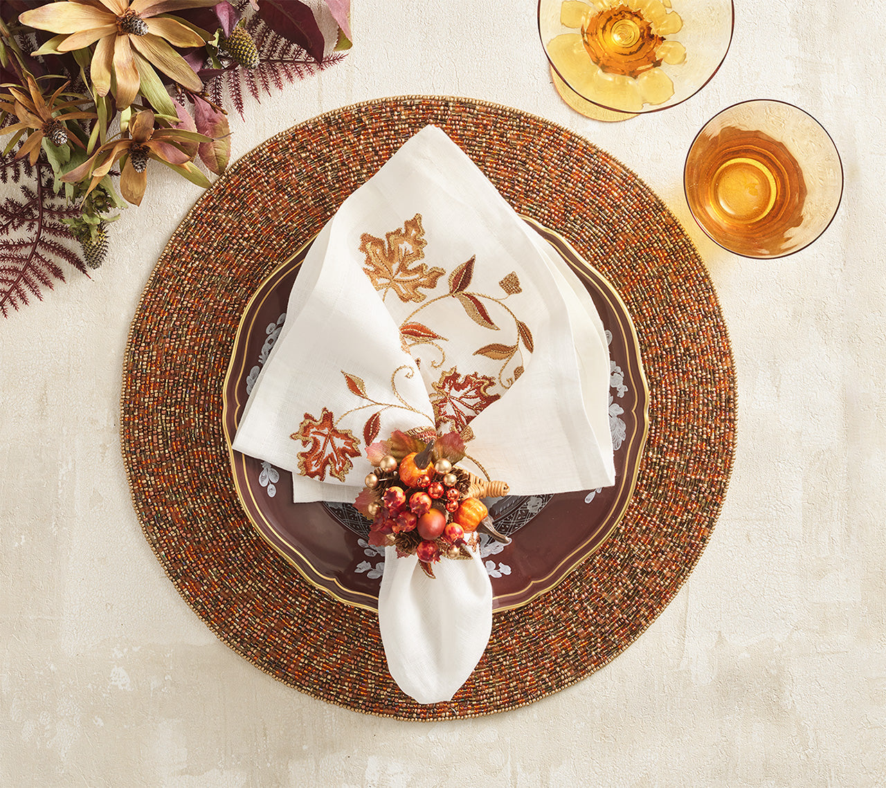 A fall-themed place setting features a beaded charger, patterned plate, embroidered napkin with a Kim Seybert Cornucopia Napkin Ring, and two amber glasses on a light surface accented with foliage.