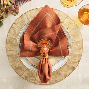 A gold-rimmed plate, rust napkin with Kim Seybert’s Turkey Trot Napkin Ring, and glasses of amber liquid on a cream table—perfect for elegant autumn decor.
