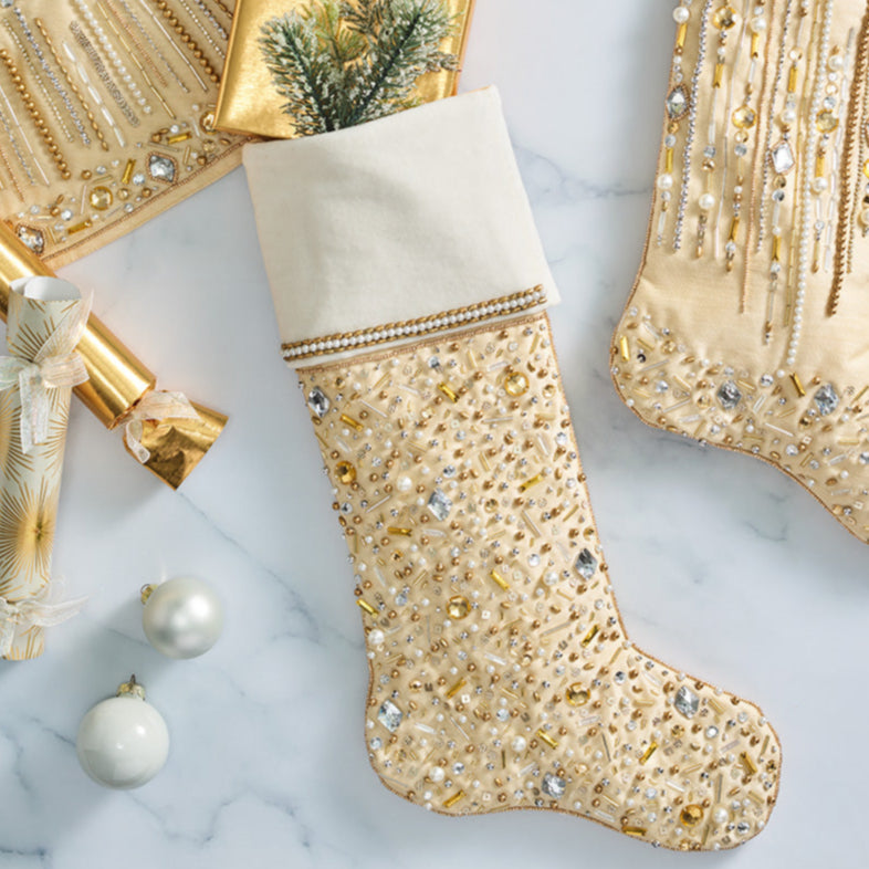 The Kim Seybert Gilded Spray Gold and Crystal Stockings, in shimmering gold and white with sequins and beads, are displayed on marble beside wrapped gifts, ornaments, and a festive holiday accent tree skirt.