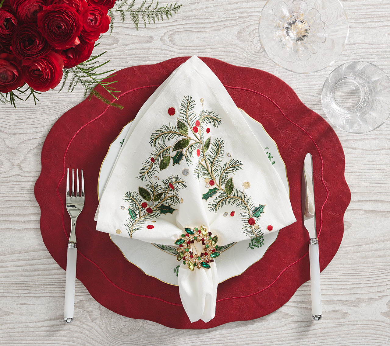 The Christmas Carol Table Linen Collection by Kim Seybert adds elegant embroidered linen detail to a holiday table set with a red placemat, white plate, festive napkin ring, utensils, wine glass, and red roses on light wood.