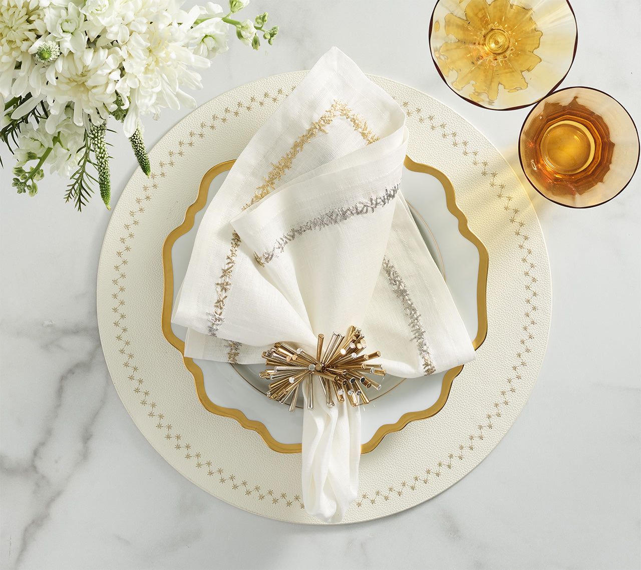 A white napkin with a gold ring sits on a white, gold-rimmed plate atop a Kim Seybert Starry Night Placemat, flanked by two amber glasses and a floral arrangement on a marble surface.