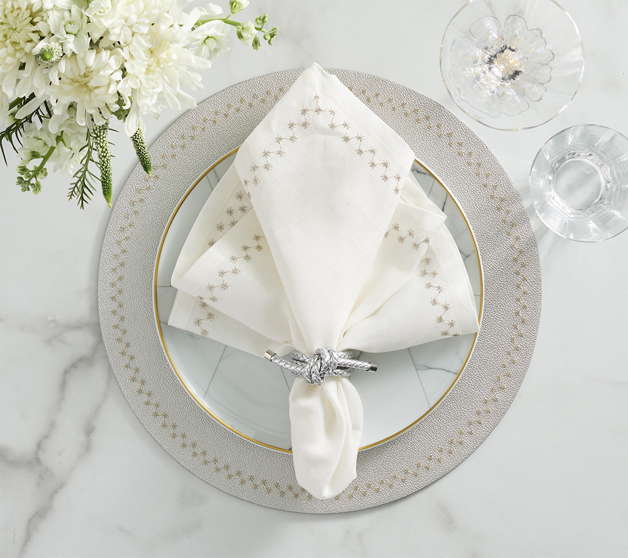 A white embroidered napkin with a silver ring rests on a decorative plate atop the Kim Seybert Starry Night Placemat, next to a glass and white flowers on a marble surface.