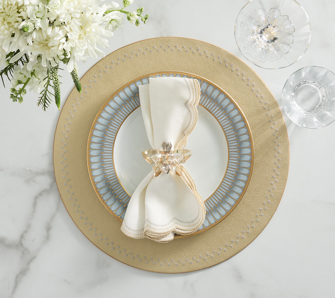 A place setting featuring the Kim Seybert Starry Night Placemat Collection, a patterned plate, white napkin with a decorative ring, and a glass of water on a white marble surface with white flowers in the upper left.