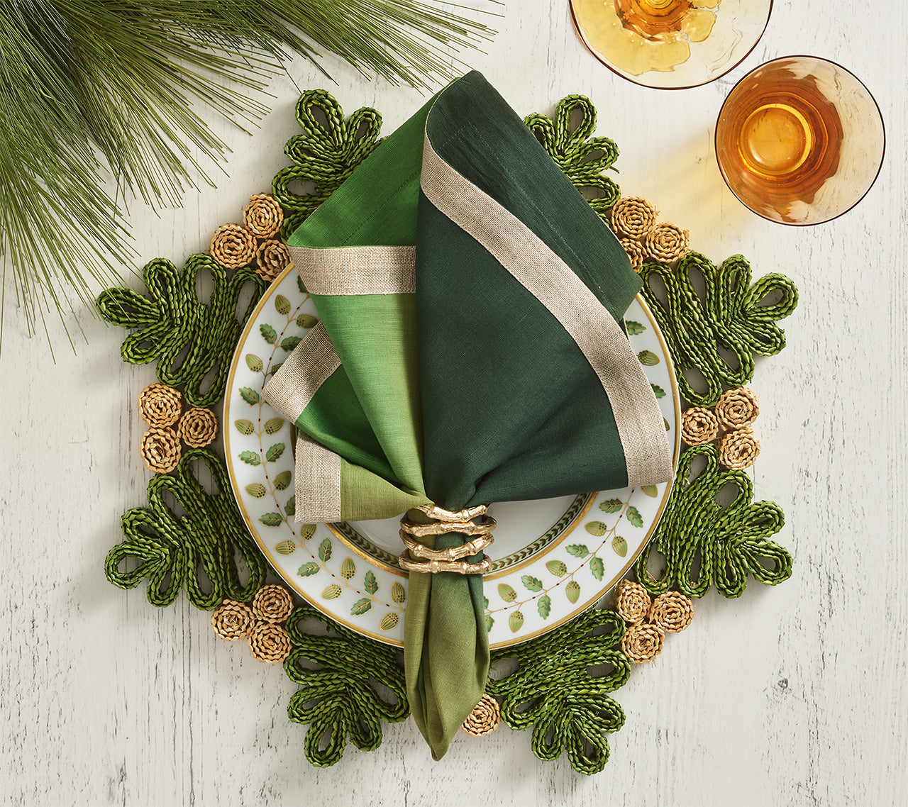 A table setting with a white plate with green leaf pattern, a green napkin and gold ring, Kim Seybert’s Verve Placemat in Green & Natural, pine needles, and two glasses filled with amber liquid.
