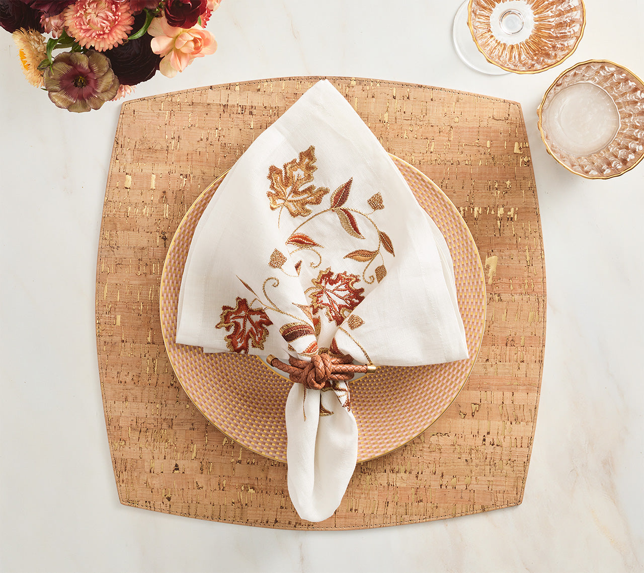 A folded white napkin with embroidered autumn leaves is held by a ring and placed on a patterned plate atop the Kim Seybert Whistler Placemat, adding rustic charm to your table. Glassware and florals complete the look.