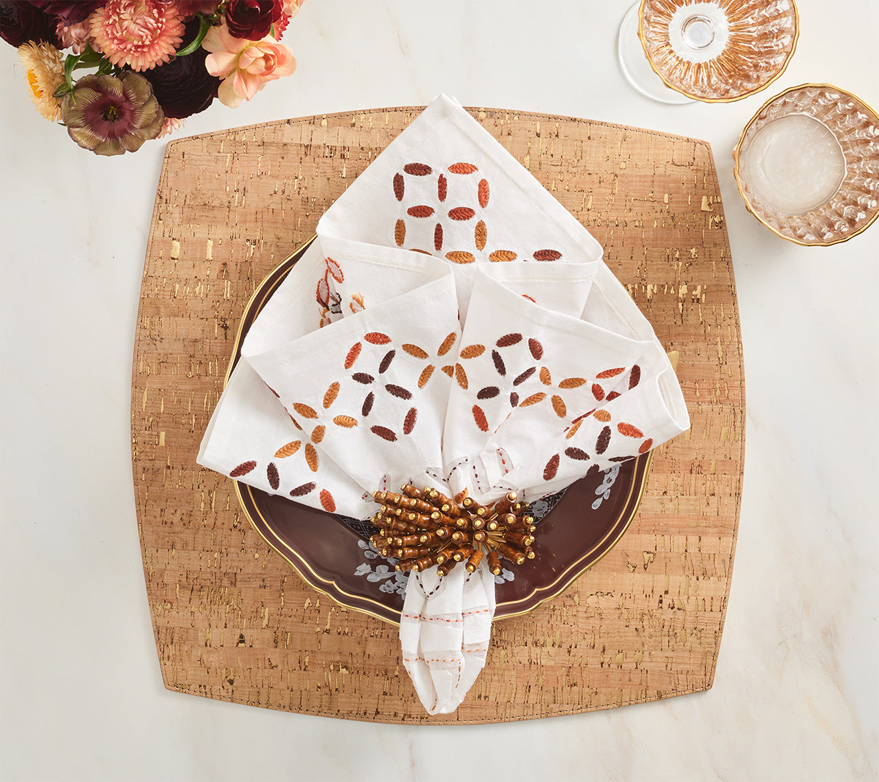 The Kim Seybert Malta Napkin Set, featuring a geometric pattern and beaded napkin ring, is styled on a gold-trimmed brown plate atop a square cork placemat, evoking the refined elegance of cutwork traditions.