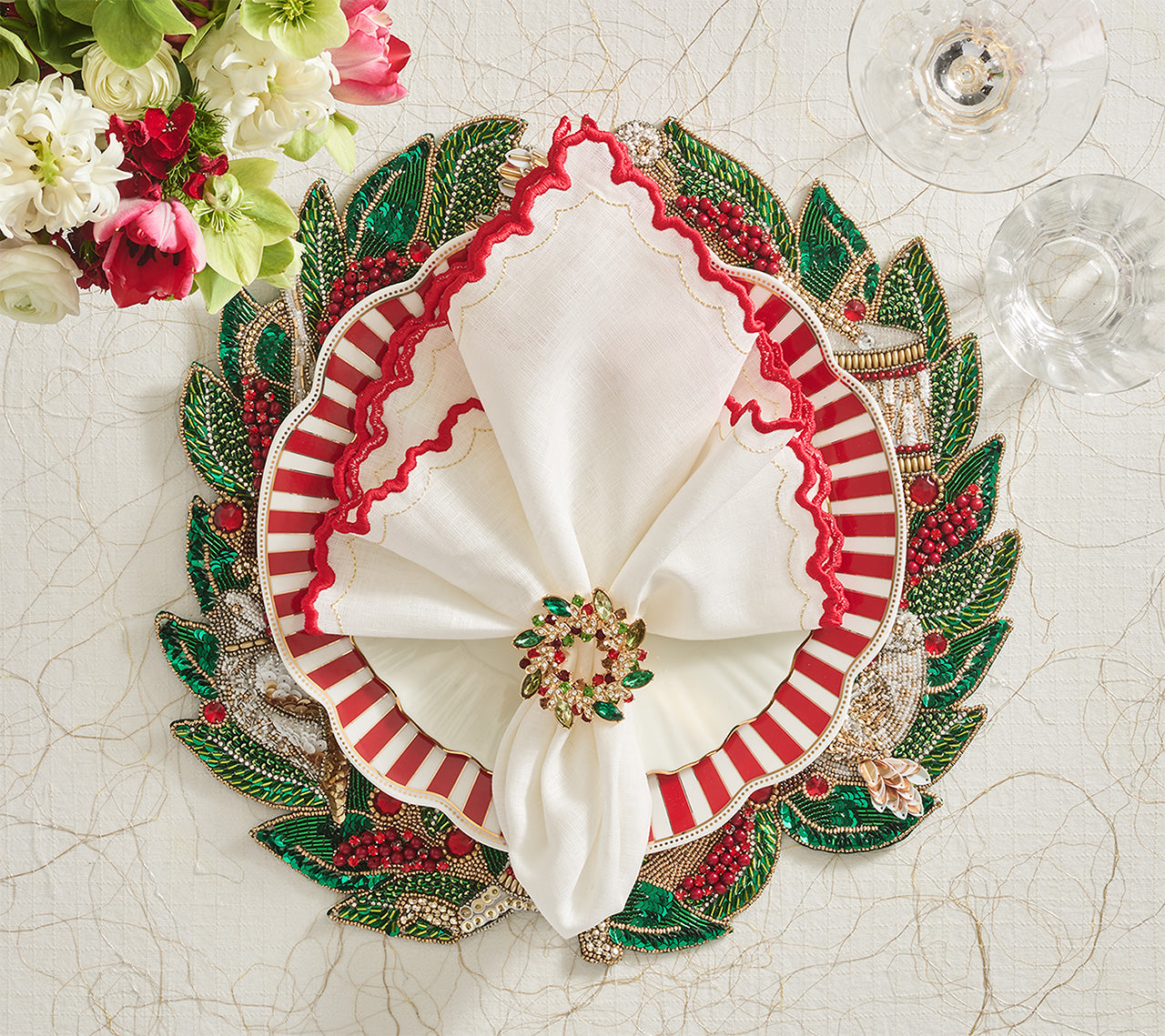 A festive table setting features a leaf charger, red and white napkins with Kim Seybert’s Gem Wreath Napkin Ring Set, two wine glasses, and fresh flowers decorating the corner.