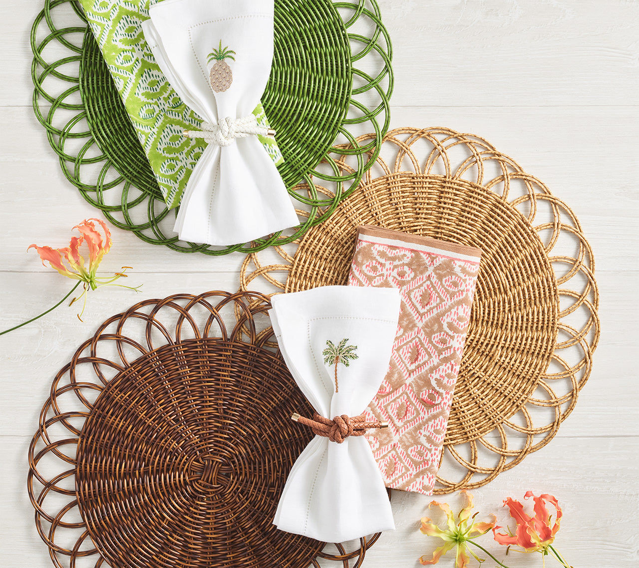 Three handwoven Alfresco Placemats by Kim Seybert, featuring scalloped edges, are arranged with patterned napkins and embroidered napkin rings on a white surface, accented by orange flowers for decoration.