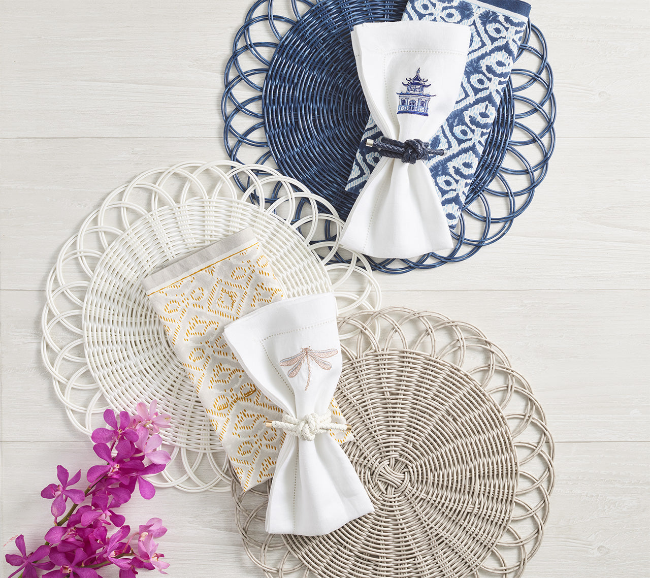 Three Kim Seybert Alfresco Placemats with scalloped edges are displayed on light wood, topped with folded napkins and rings, handwoven faux rattan textures, and purple flowers accenting the lower left corner.