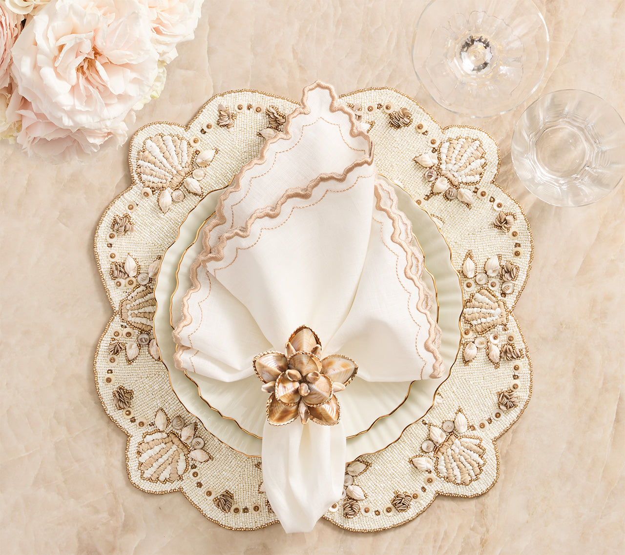 A white plate with a folded napkin in the Kim Seybert Beachcomber Napkin Ring Set rests on a cream, scalloped placemat with shell and bead accents, beside clear glasses and blush flowers—ideal for coastal table decor.