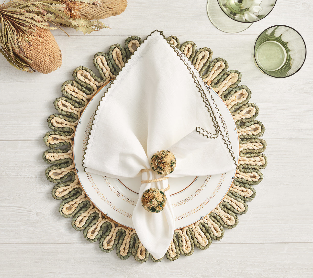 A white napkin in a Kim Seybert Casbah Napkin Ring sits on a decorative plate and placemat, complemented by dried flowers, two green glasses, and organic raffia accents for an elegant table setting.