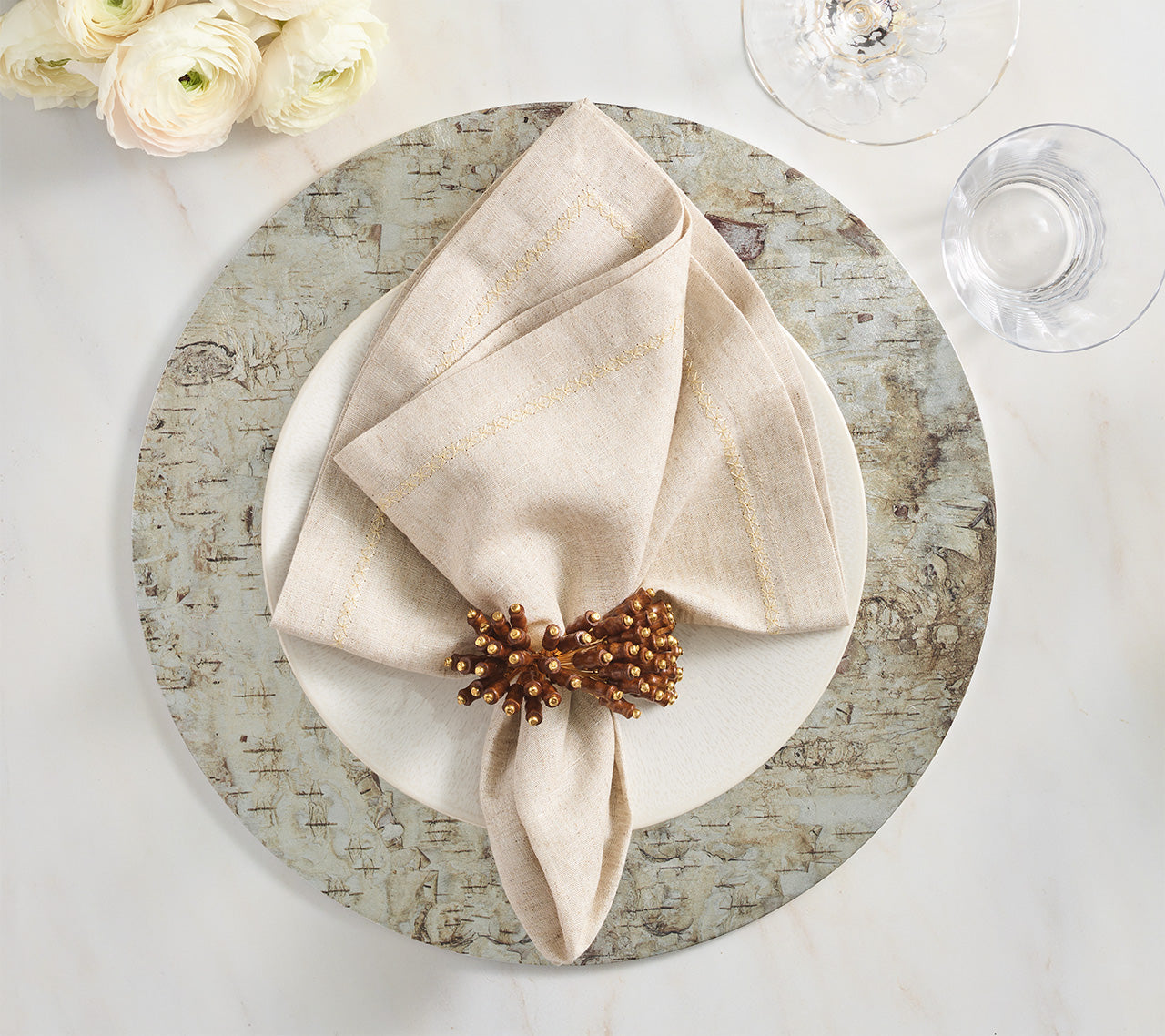 A beige napkin with a brown ring sits on a white plate and the Kim Seybert Chalet Placemat, creating nature-inspired table decor beside glasses and white flowers.