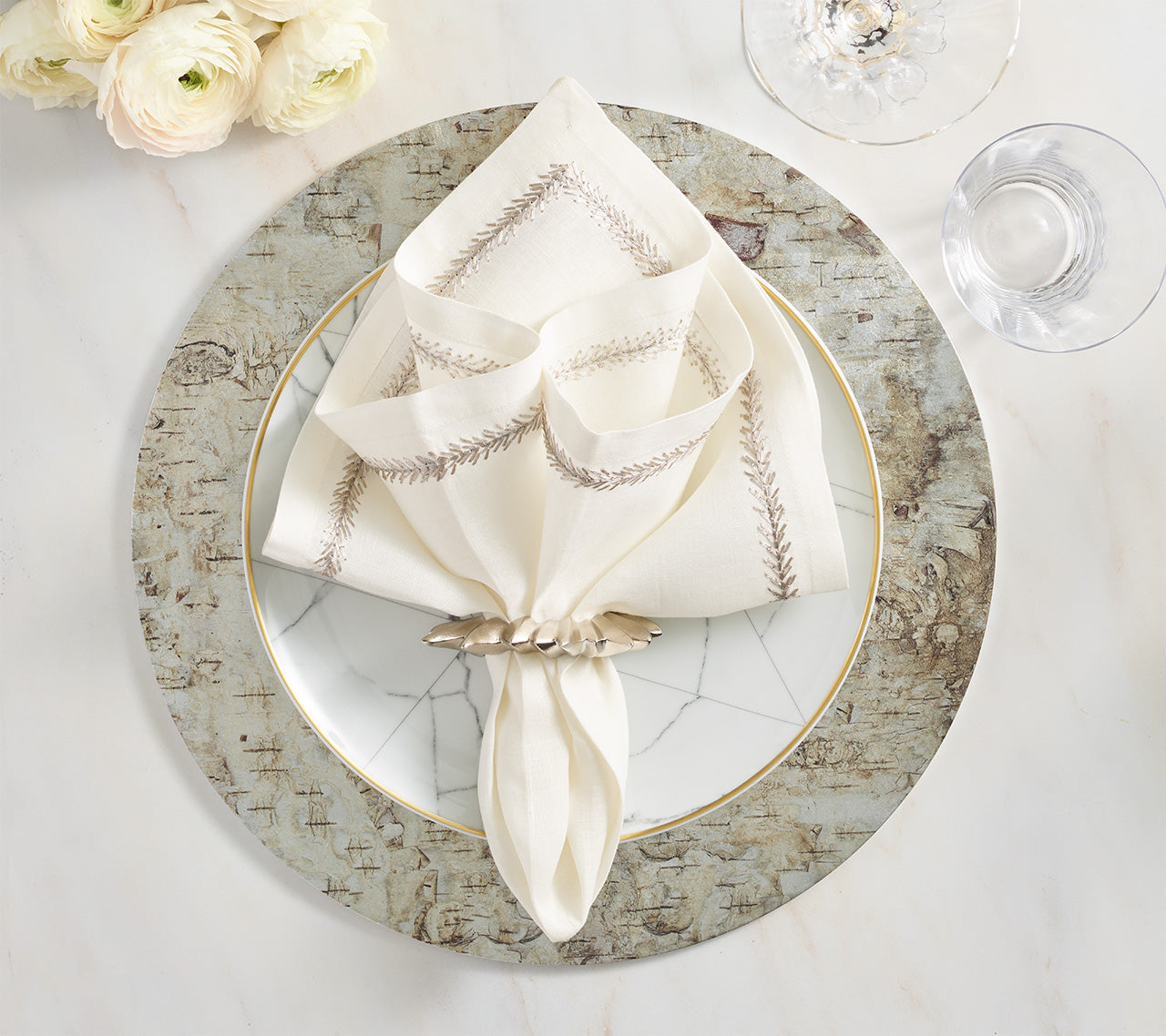 A folded embroidered napkin with a napkin ring rests on a white plate with a silver rim, set atop the Kim Seybert Chalet Placemat, next to a wine glass and white flowers for a nature-inspired table setting.