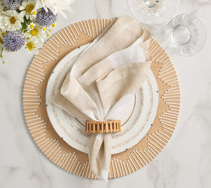A beige napkin with a Kim Seybert Spoke Napkin Ring rests on layered white plates with dotted edges atop a textured placemat; two empty glasses and a floral arrangement complete the elegant table decor.